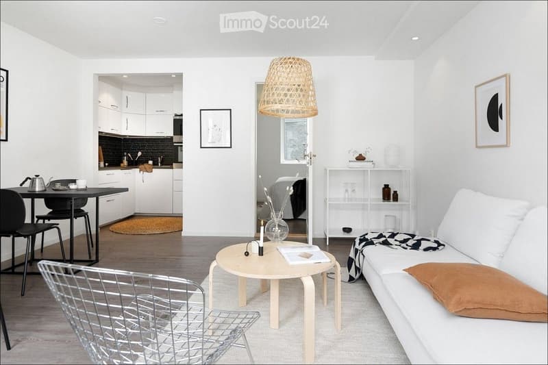 Modern, minimalist living room with white walls, light wood flooring, and a white sofa with a brown throw pillow. A round wooden coffee table holds a candle and a vase with dried flowers. A metal wire chair is near the coffee table. The room features a sm