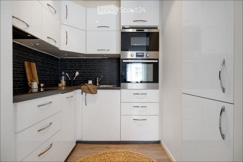 Modern kitchen with white cabinets, stainless steel oven, and black tiled backsplash. Includes a wooden cutting board, cup, and various kitchen utensils on the counter.