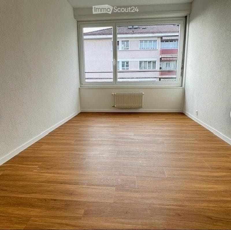 Empty room with wooden flooring, white walls, and a radiator under a large window overlooking a building.