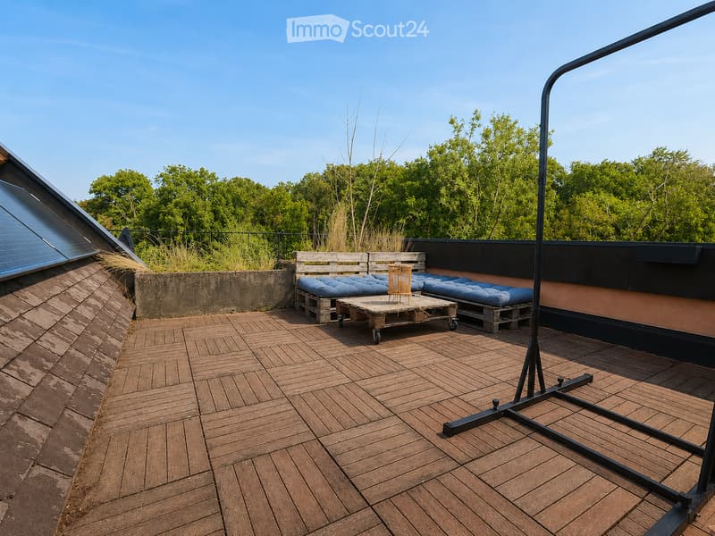Wooden deck with benches, a small table, and a chair, surrounded by greenery and a clear blue sky.