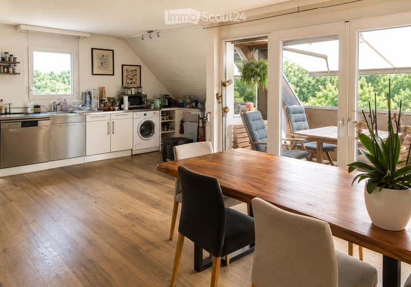 Bright, open-plan kitchen and dining area with wooden flooring, white cabinets, stainless steel appliances, and a large dining table with chairs. A balcony with outdoor seating is visible through large glass doors.