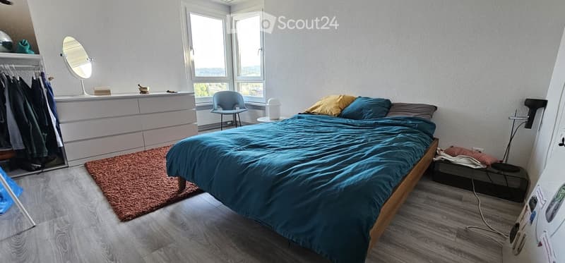 Cozy bedroom with a wooden bed, blue duvet, a small chair, a mirror, a dresser, and a clothing rack. The room has large windows with a scenic view and a warm, inviting atmosphere.