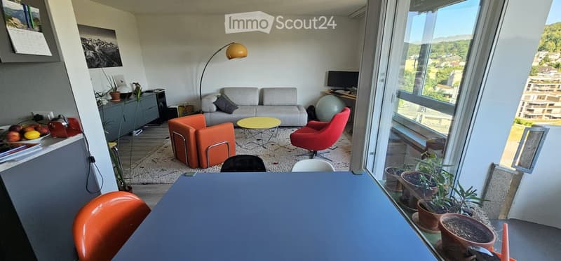 Modern living room with large windows offering a scenic view, featuring a grey sofa, orange armchairs, a yellow coffee table, a floor lamp, and a screen on a desk.