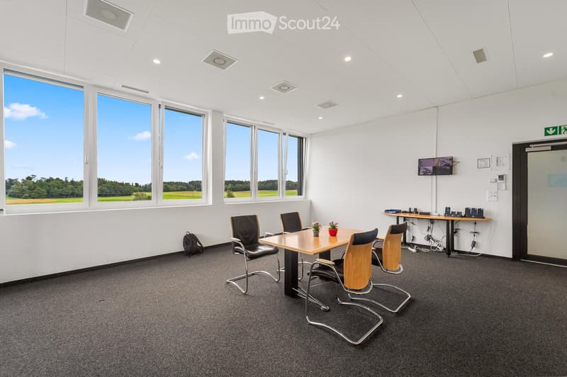 Bright, modern meeting room with large windows, a wooden table, black chairs, a screen, and a backpack on the floor.