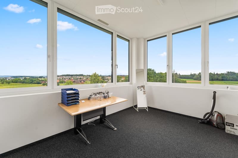 Bright, unfurnished room with large windows offering a scenic view, a small table with office supplies, a mirror, and a vacuum cleaner in the corner.