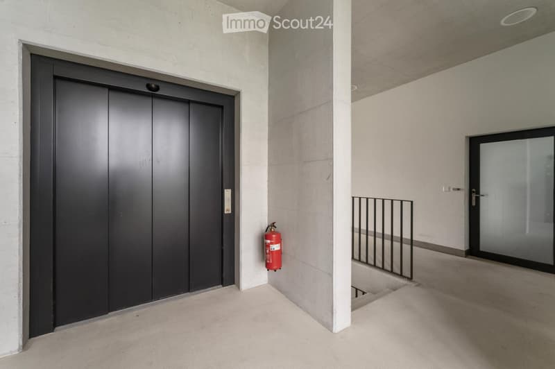 Modern hallway with a sleek black elevator door, a red fire extinguisher mounted on the wall, a white pillar, and a small staircase leading upwards with a black railing.