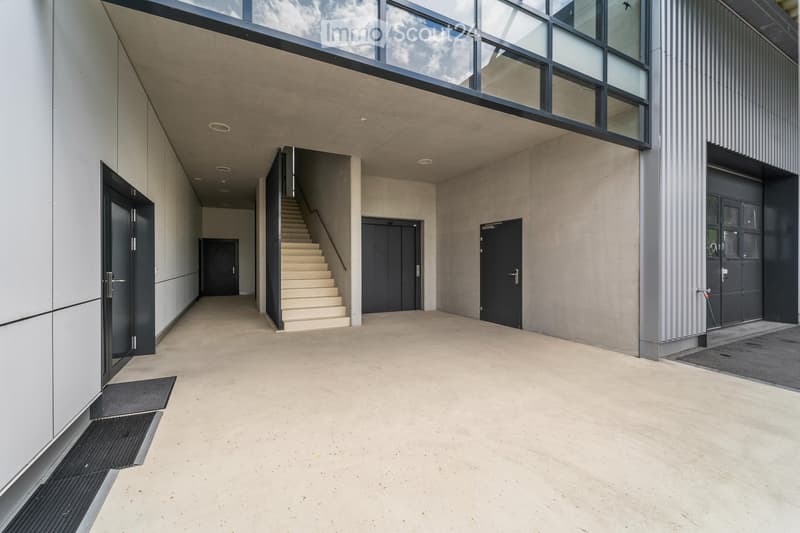 Modern, minimalist hallway with concrete floors, grey walls, and a staircase leading upwards. Features include recessed lighting, black doors, and a sleek, contemporary design.