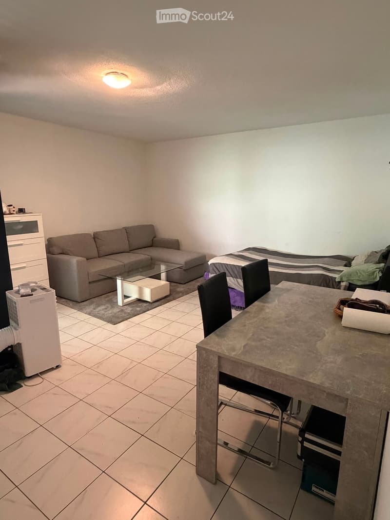 A modern, multi-functional living space with a sofa, coffee table, dining table with chairs, a bed, and an air conditioner. The room features tiled flooring and white walls.