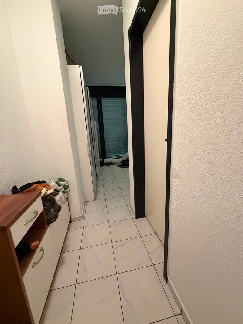 A narrow hallway with tiled flooring, white walls, and a wooden shoe rack on the left side. The hallway leads to an open doorway at the end.