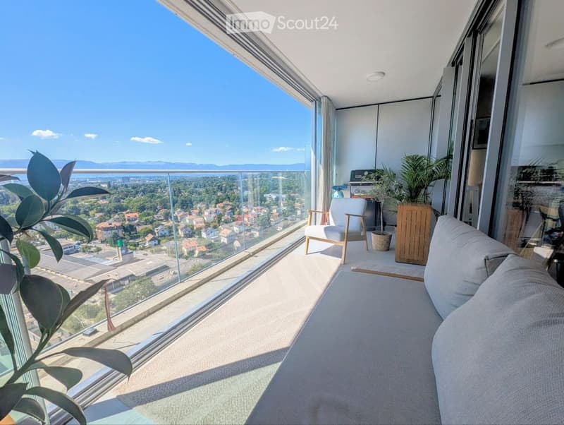 Modern balcony with large glass windows, offering a panoramic city view. Features a grey sofa, wooden chair, small table, and potted plants.