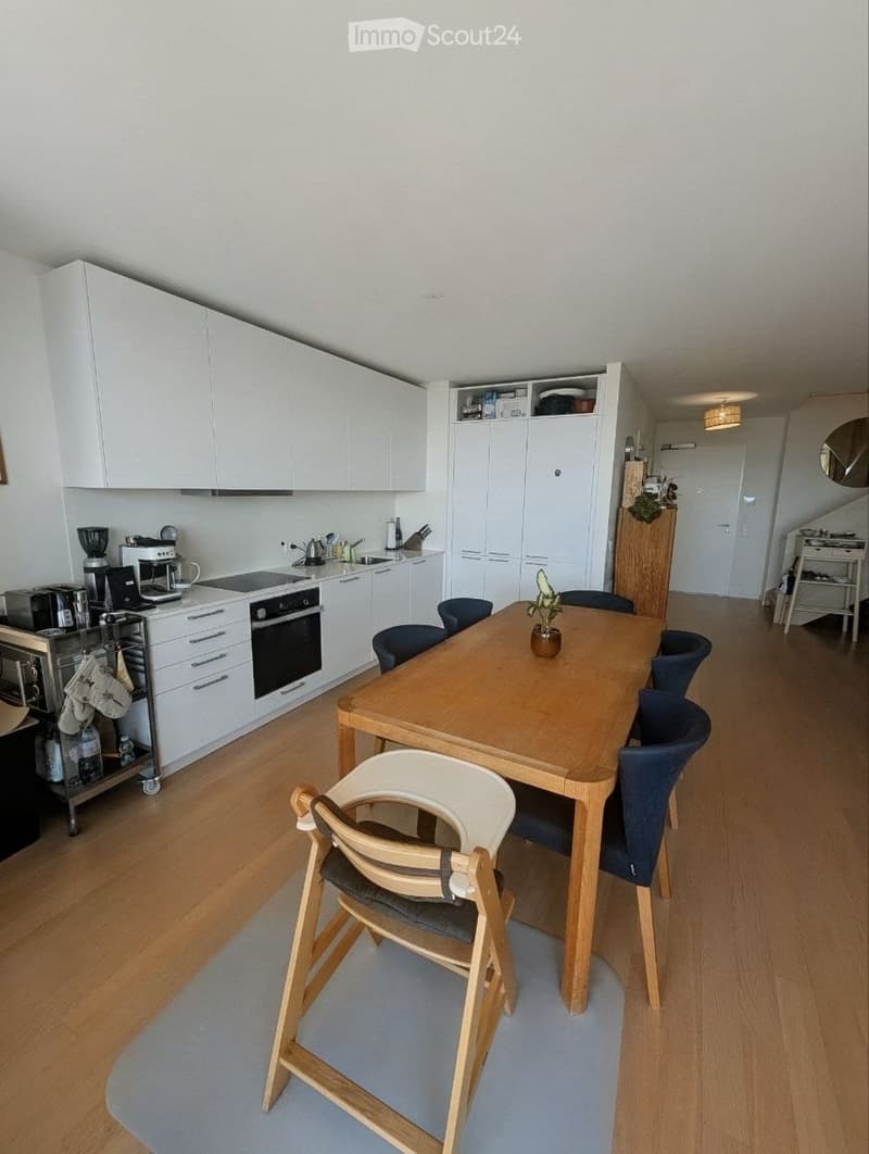 Modern kitchen with white cabinets, wooden dining table, blue chairs, and various appliances including a coffee maker, toaster, and microwave. A baby chair is also present near the table.