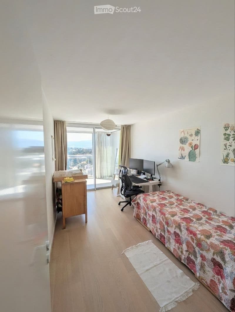 Bright room with wooden flooring, large window with view, desk with computer, chair, bed with floral cover, rug, and cabinet.