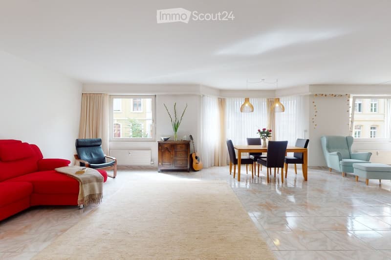Bright living and dining area with a red sofa, a light blue armchair, a wooden dining table with chairs, a guitar, and a cabinet with a flower vase. Natural light streams in through large windows with curtains.