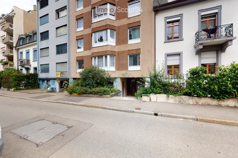 Multi-story residential building with multiple windows, balconies, and greenery in front.