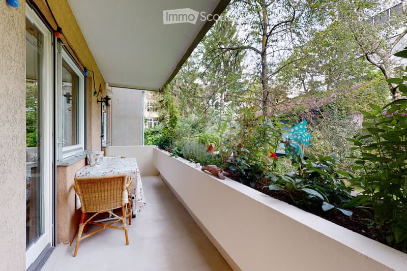 Balcony with a small table and a wicker chair, surrounded by lush greenery and plants, with a view of trees and neighboring buildings.
