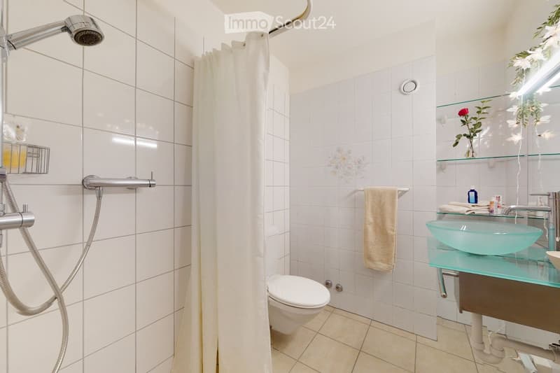 Modern bathroom with white tiles, a glass sink, a toilet, a shower with a handheld sprayer, and a towel on a rack.