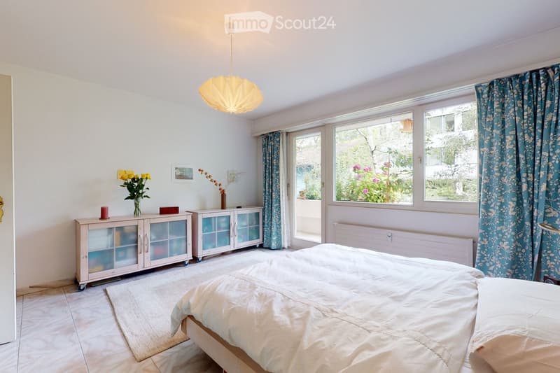 Bright bedroom with a large bed, white walls, large windows with floral curtains, a cabinet with glass doors, a vase with flowers, and a hanging lamp.