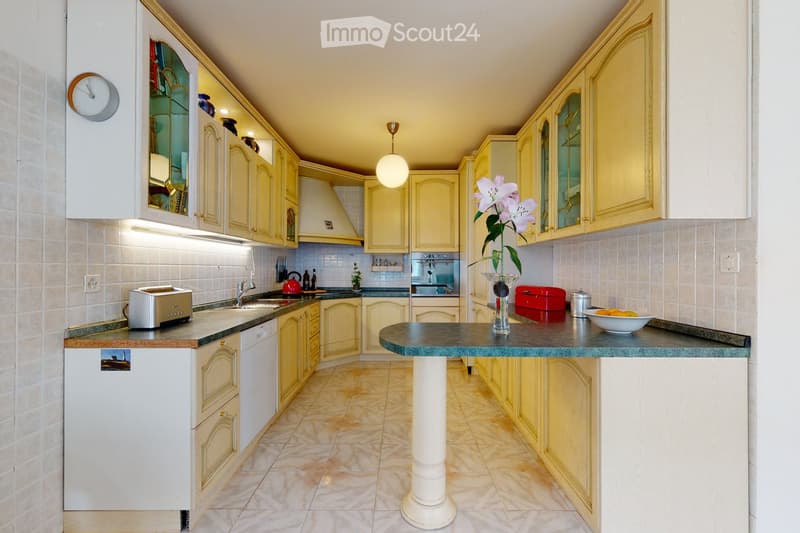 Bright kitchen with yellow cabinets, white tiled walls, and a central island with a vase of lilies. Appliances include a refrigerator, oven, and toaster, with a sink and faucet on the left side.