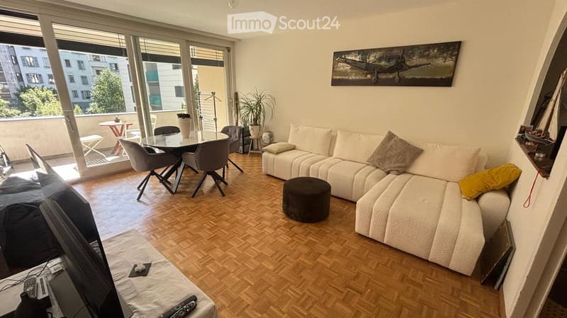 Modern living room with a white sectional sofa, ottoman, coffee table, dining table, chairs, and a balcony with outdoor seating.