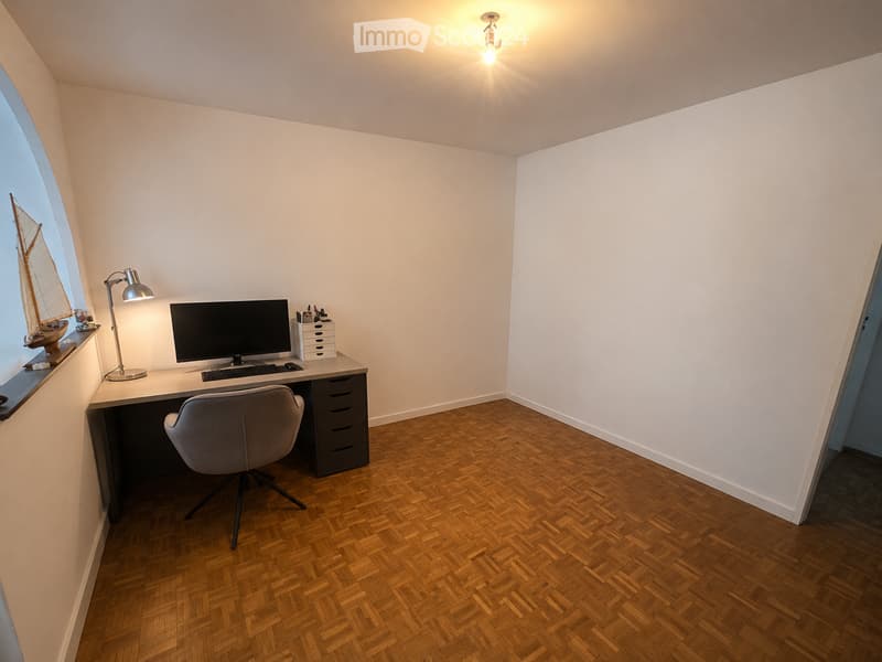 A modern, well-lit study or home office with a wooden herringbone floor, white walls, and a large window. The room features a sleek desk with a computer setup, a comfortable office chair, a desk lamp, and a decorative model ship on the windowsill.