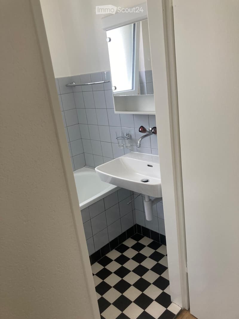 Small bathroom with a bathtub, white sink, mirror, and tiled walls. The floor features a black and white checkered pattern.