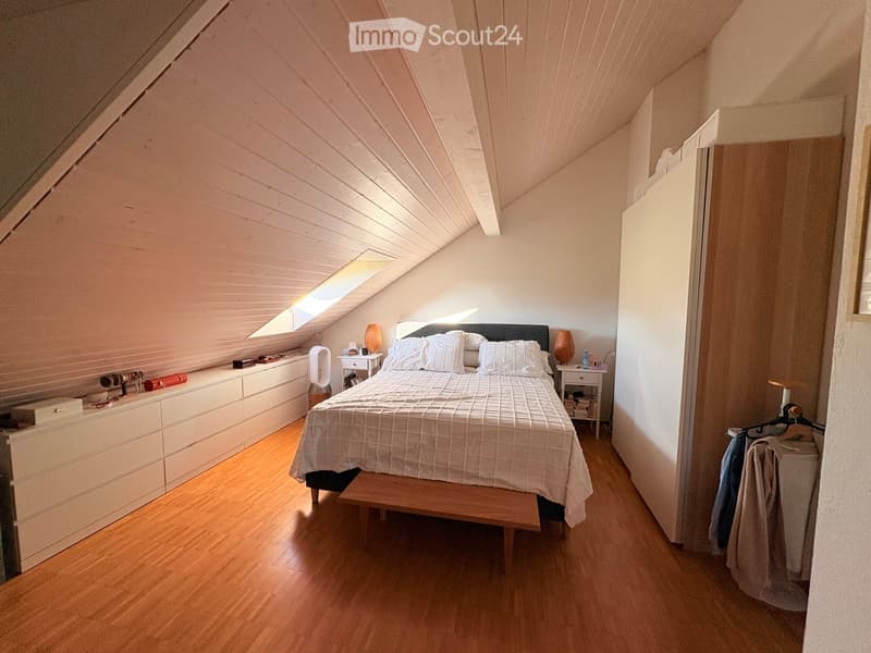 Cozy attic bedroom with wooden floors, slanted wooden ceiling, skylight window, bed with white bedding, nightstands, lamps, and a small fan. Features built-in storage and a clothing rack.