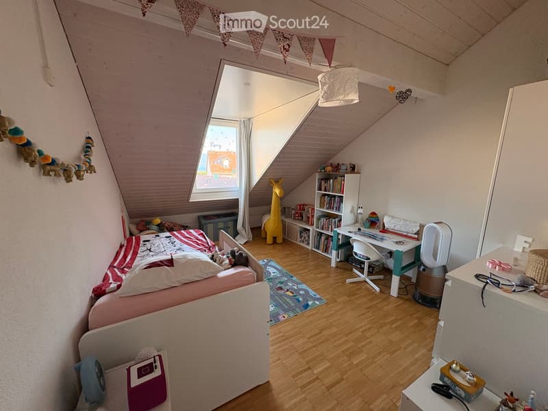 Cozy children's bedroom with slanted wooden ceiling, large window, bed with colorful bedding, desk with chair, bookshelf, and various toys and decorations.