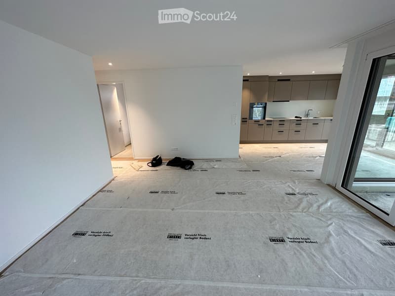 Empty room with white walls, covered floor, and a partially visible kitchen area with cabinets and appliances in the background.