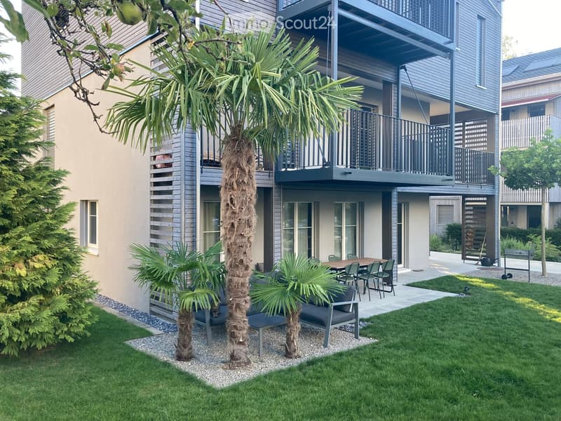 Well-maintained garden area with palm trees, a dining table with chairs, and a modern building with balconies in the background.