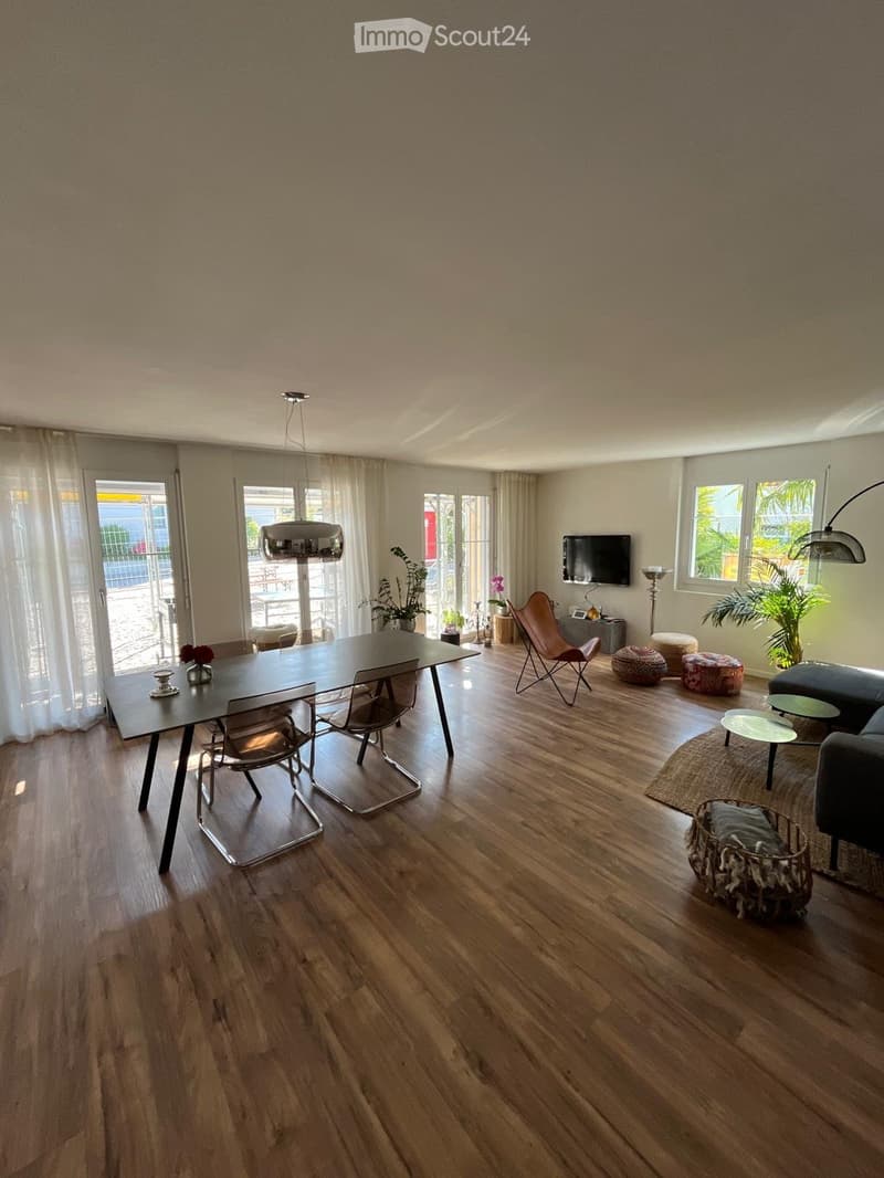 Open-plan living and dining area with wooden flooring, large windows, and modern furniture including a sofa, dining table, chairs, and decorative plants.