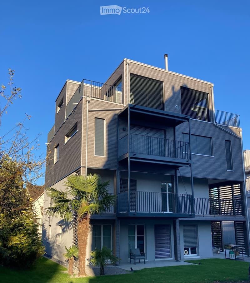 Modern two-story house with grey wooden exterior, multiple balconies, large windows, and a small garden area with palm trees and a bench.