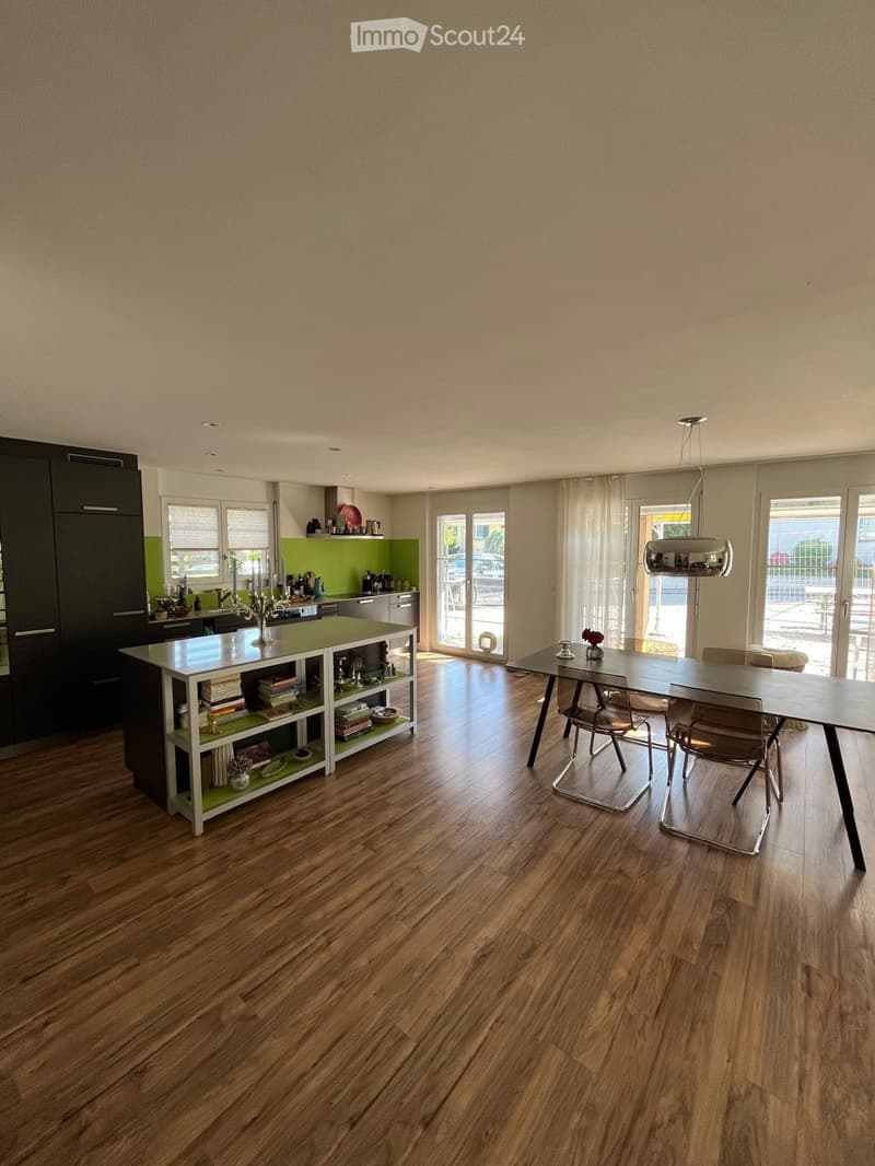 Open-plan kitchen and dining area with wooden flooring, modern kitchen island, dining table, and chairs. Features include large windows with curtains, a hanging pendant light, and various kitchen items on the island and shelves.