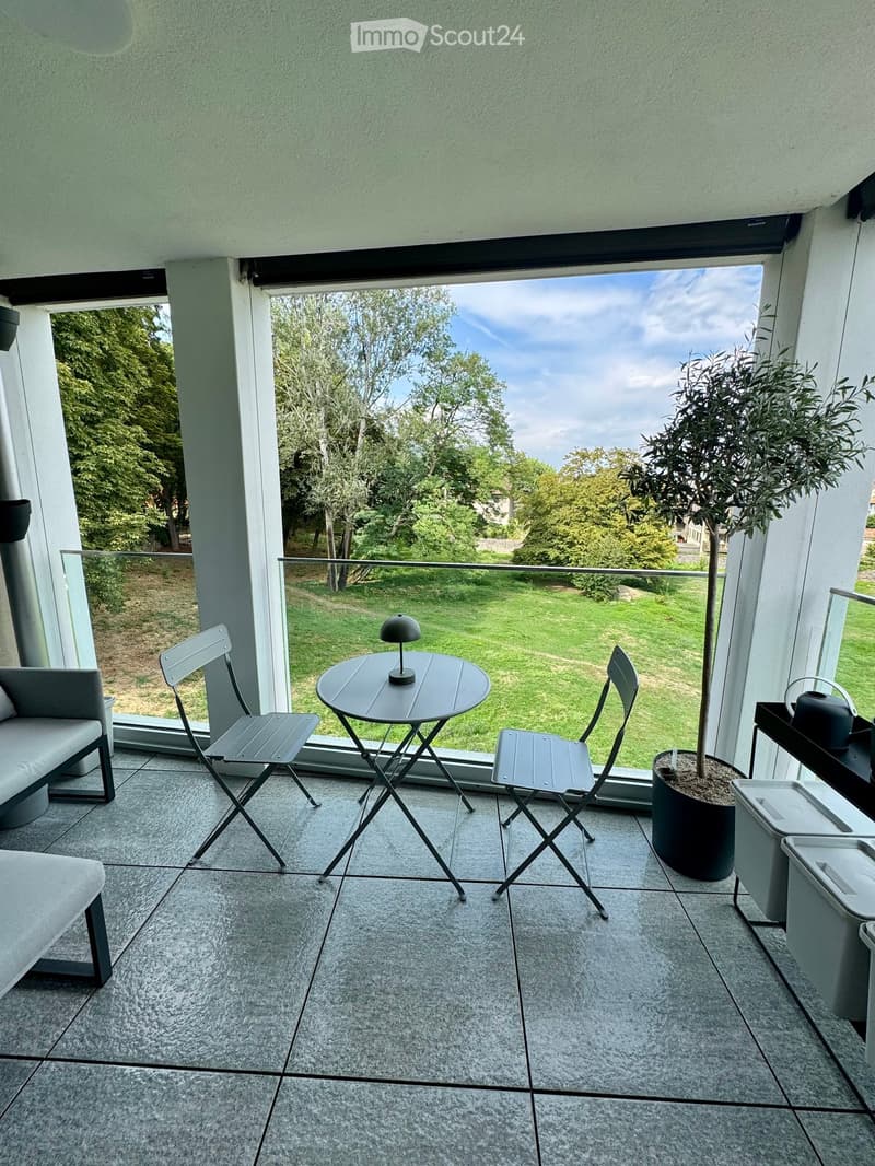 Modern balcony with grey tiled floor, glass railing, grey folding chairs, small round table with lamp, grey sofa, potted tree, and scenic view of green lawn and trees.