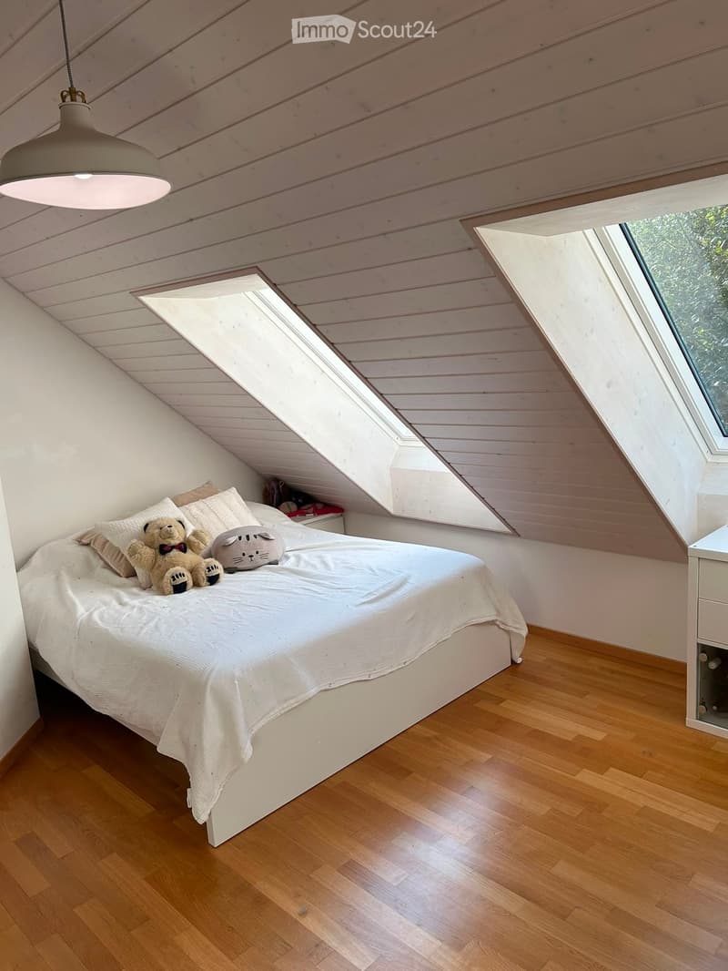 Cozy attic bedroom with wooden floors, slanted wooden ceiling, skylights, and a bed with pillows and stuffed animals.