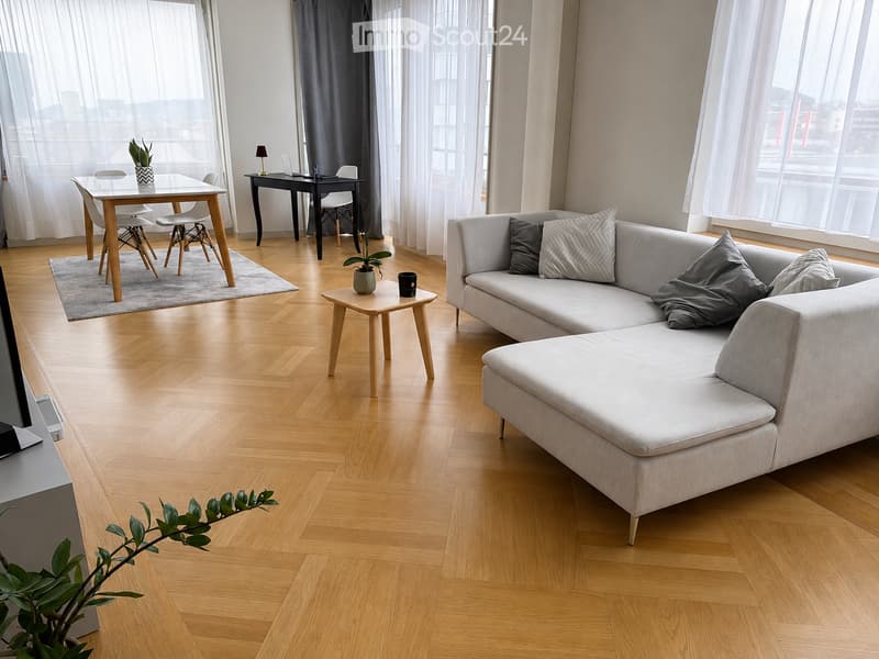 Modern living room with light wooden flooring, large windows with sheer curtains, grey sectional sofa, coffee table, side table, dining table with chairs, and a small desk with a laptop.