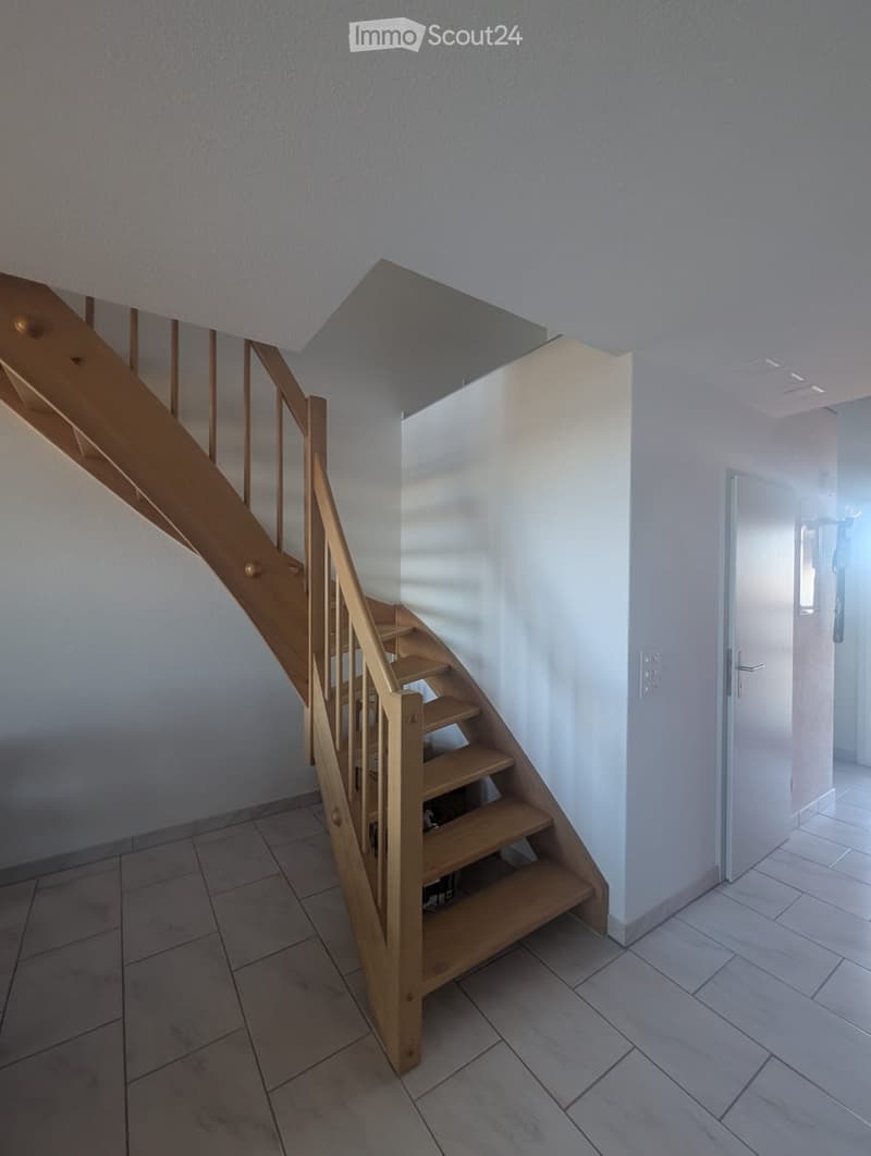 Wooden staircase with a handrail, leading to an upper level, with tiled floor and white walls.