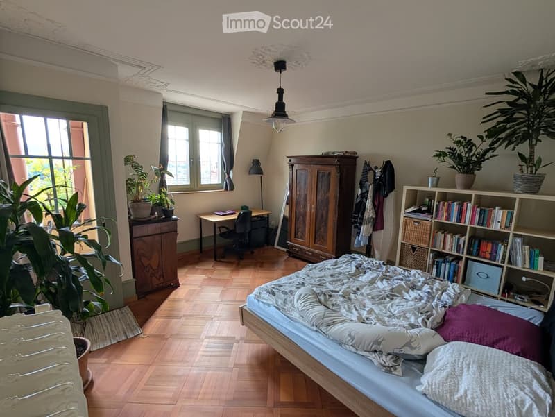 Cozy bedroom with wooden flooring, large bed, bookshelf filled with books, desk with chair, wardrobe, and several potted plants near the windows.