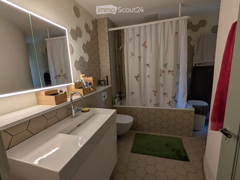 Modern bathroom with hexagonal tiles, double sink, large mirror with LED lighting, shower area with bird-patterned curtain, toilet, and a green bath mat.