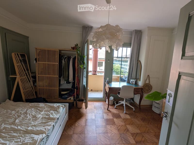 Cozy bedroom with a bed, desk, chair, wardrobe, and a balcony view. Features wooden flooring, a hanging lamp, and a potted plant.