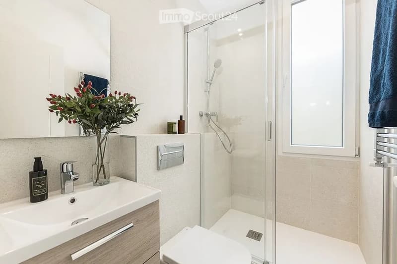Modern bathroom with a white sink, chrome faucet, large mirror, glass-enclosed shower, and a towel rack. A vase with flowers and a bottle are placed on the sink counter.
