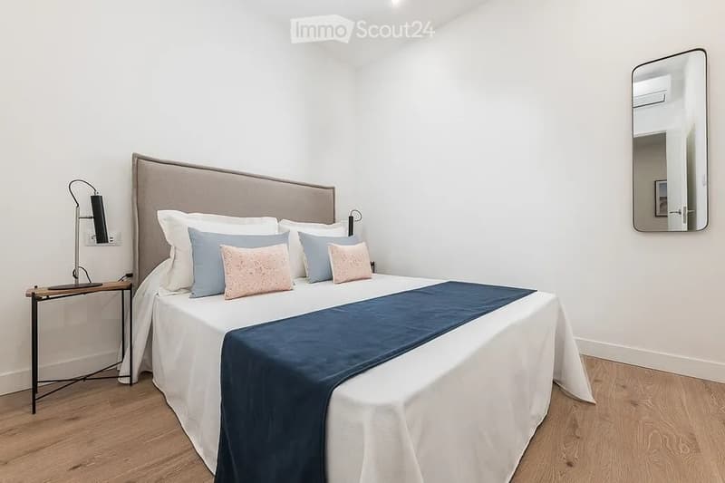 Modern bedroom with a large bed, white bedding, decorative pillows, a nightstand with a lamp, and a large mirror on the wall.