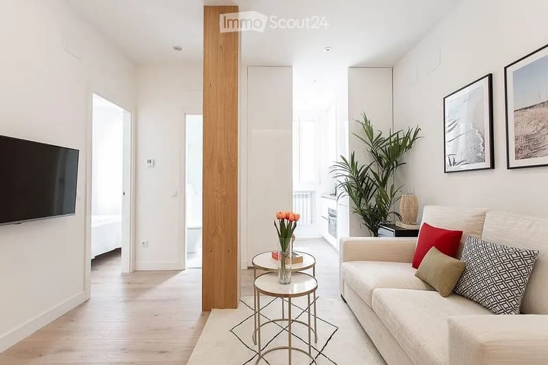 Modern living room with light-colored walls, wooden flooring, a white sofa with colorful cushions, a coffee table, a small round table with a vase of flowers, and a potted plant near the window.