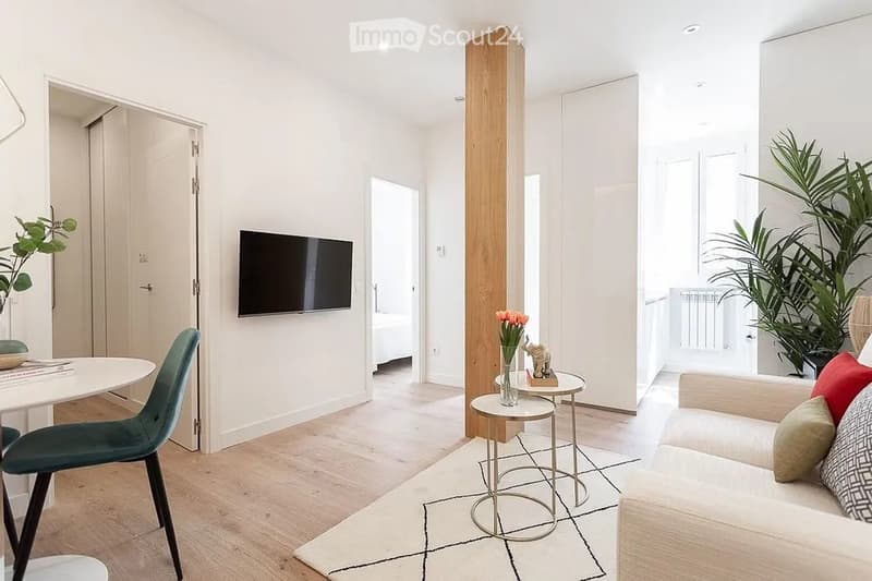 Modern living room with white walls, light wood flooring, and large windows. Features a white sofa with colorful cushions, a coffee table, a dining table with chairs, a television mounted on the wall, and decorative plants.