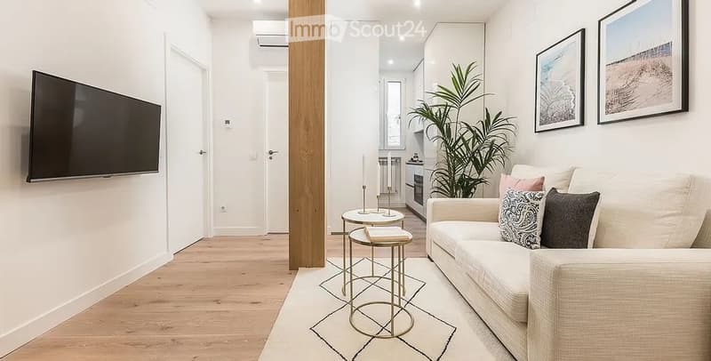 Modern living room with light-colored sofa, coffee table, small round stools, a potted plant, and a flat-screen TV mounted on the wall. The space features light wood flooring and white walls.