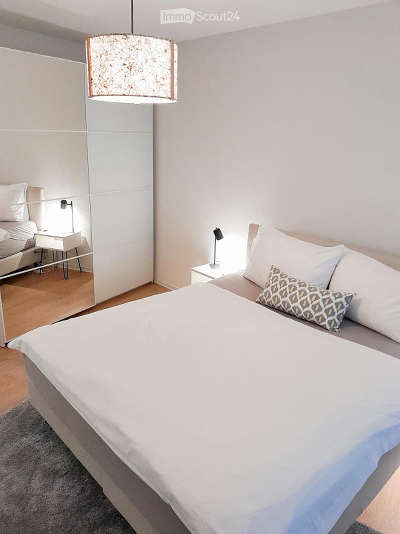Modern bedroom with a large bed, white bedding, decorative pillows, nightstands with lamps, and a large mirrored wardrobe.