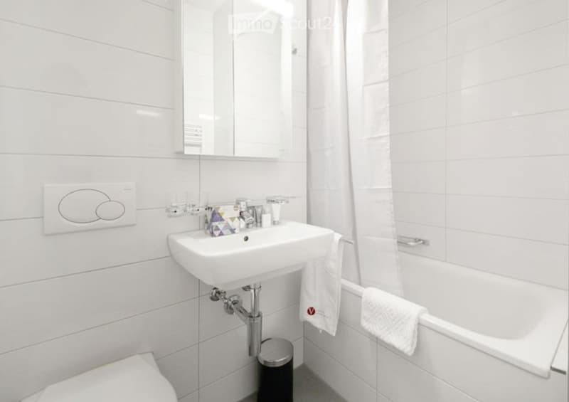Modern bathroom with white tiles, a sink, a mirror, a bathtub, and a toilet. White towels are neatly placed on the edge of the bathtub.