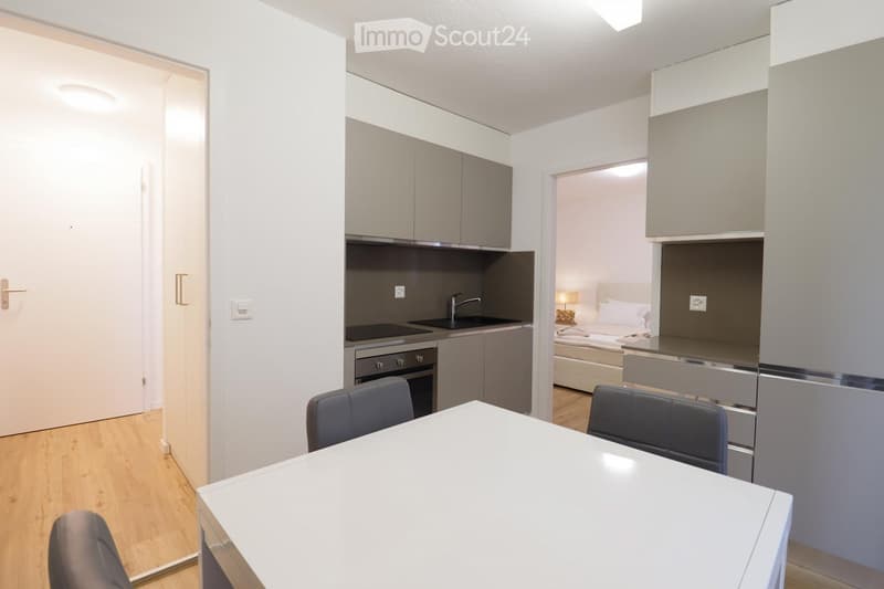 Modern, compact studio apartment with a combined living, dining, and sleeping area. Features a white dining table with chairs, a small kitchen area with cabinets, sink, and oven, and a bed with a lamp in the sleeping area.