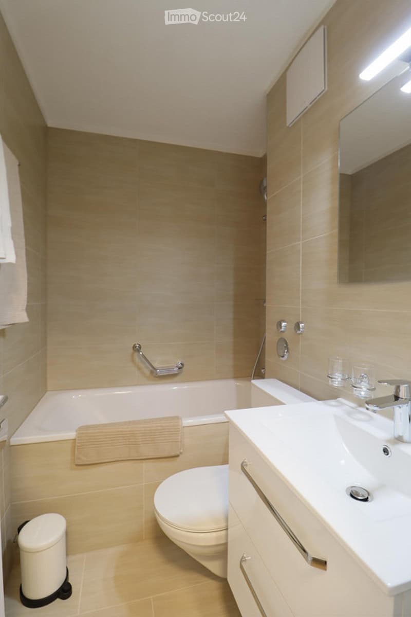 Modern bathroom with beige tiles, a bathtub, toilet, sink, and a mirror above the sink. Two cups are placed on the sink counter, and a small trash can is near the toilet.