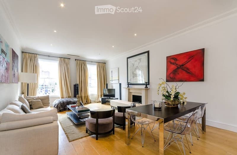 Modern living room with large windows, fireplace, sofa, coffee table, armchairs, ottoman, floor lamp, TV, and dining area with a table and chairs.