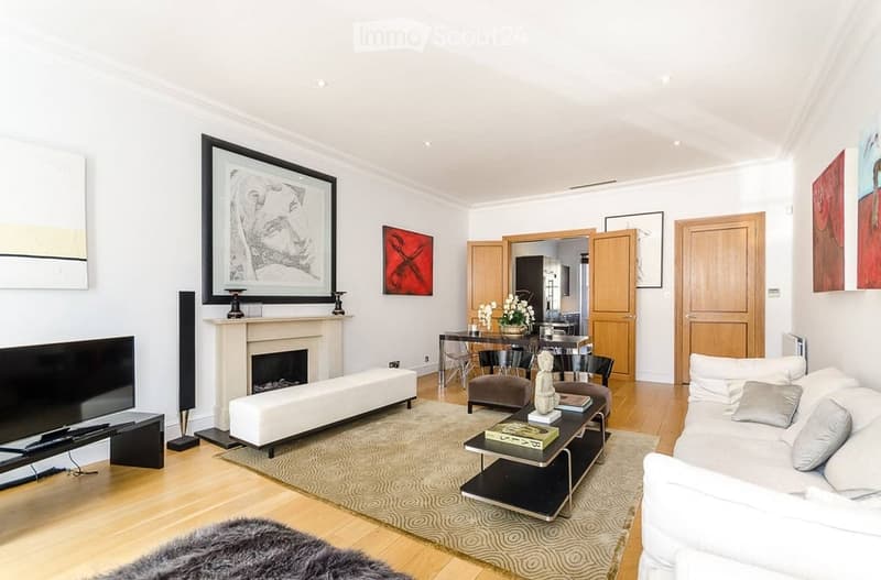 Modern living room with a white sofa, coffee table, fireplace, TV, and artwork on the walls. The space features wooden flooring and a dining area visible in the background.